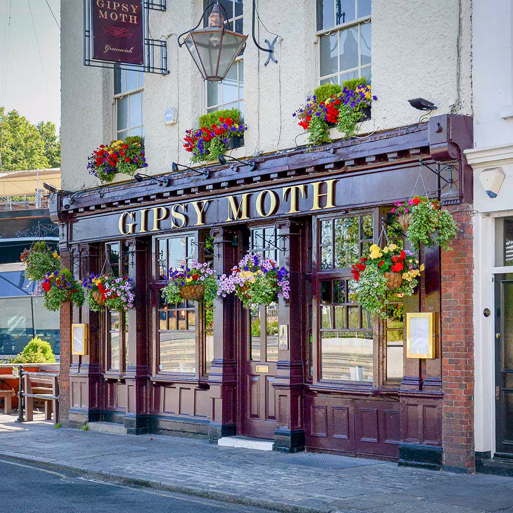 The traditional street-front facade of the refurbished Gipsy Moth pub, showing the dark wood panelling, gold lettering, and vibrant hanging flower baskets.