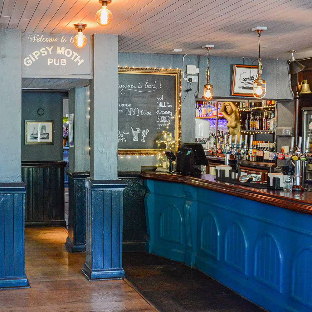 The refurbished bar area of the Gipsy Moth pub, showing the blue-painted bar, beer taps, a welcome chalkboard, and warm, industrial-style lighting.