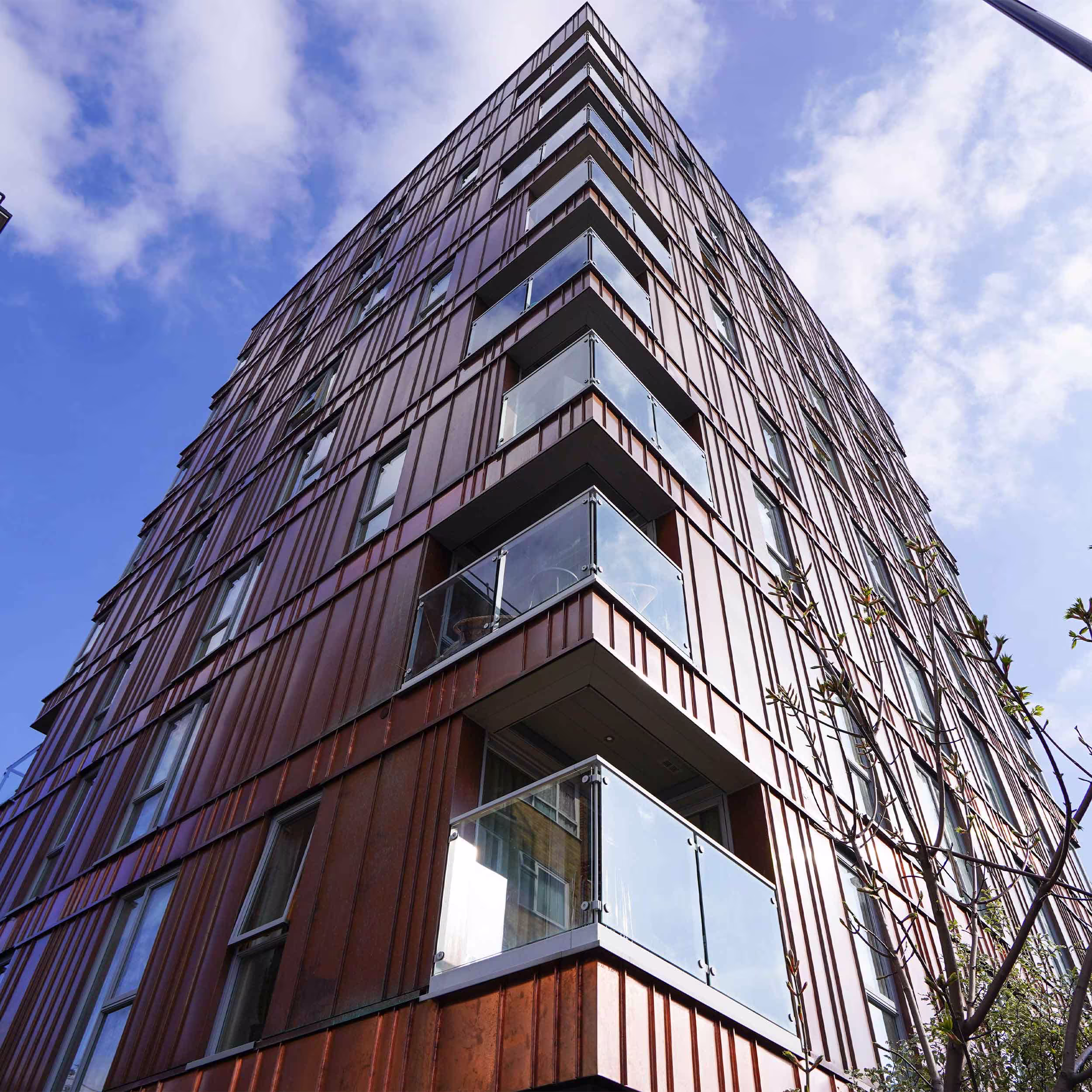 A dramatic low-angle shot looking up the corner of a tall, modern residential building, highlighting its new copper-coloured vertical panel cladding and glass balconies.