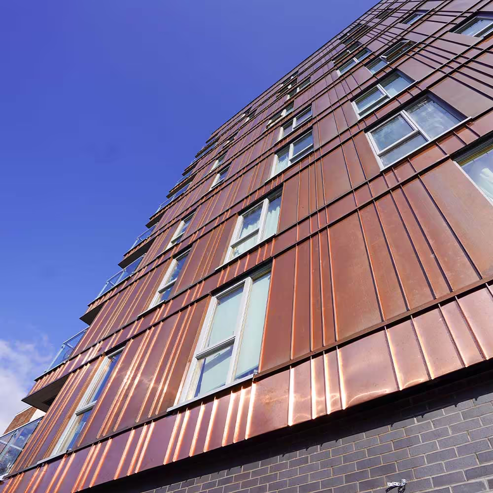 Looking up the facade of a re-cladded tower block, showing the detail and texture of the new copper-coloured standing seam metal cladding against a bright blue sky.