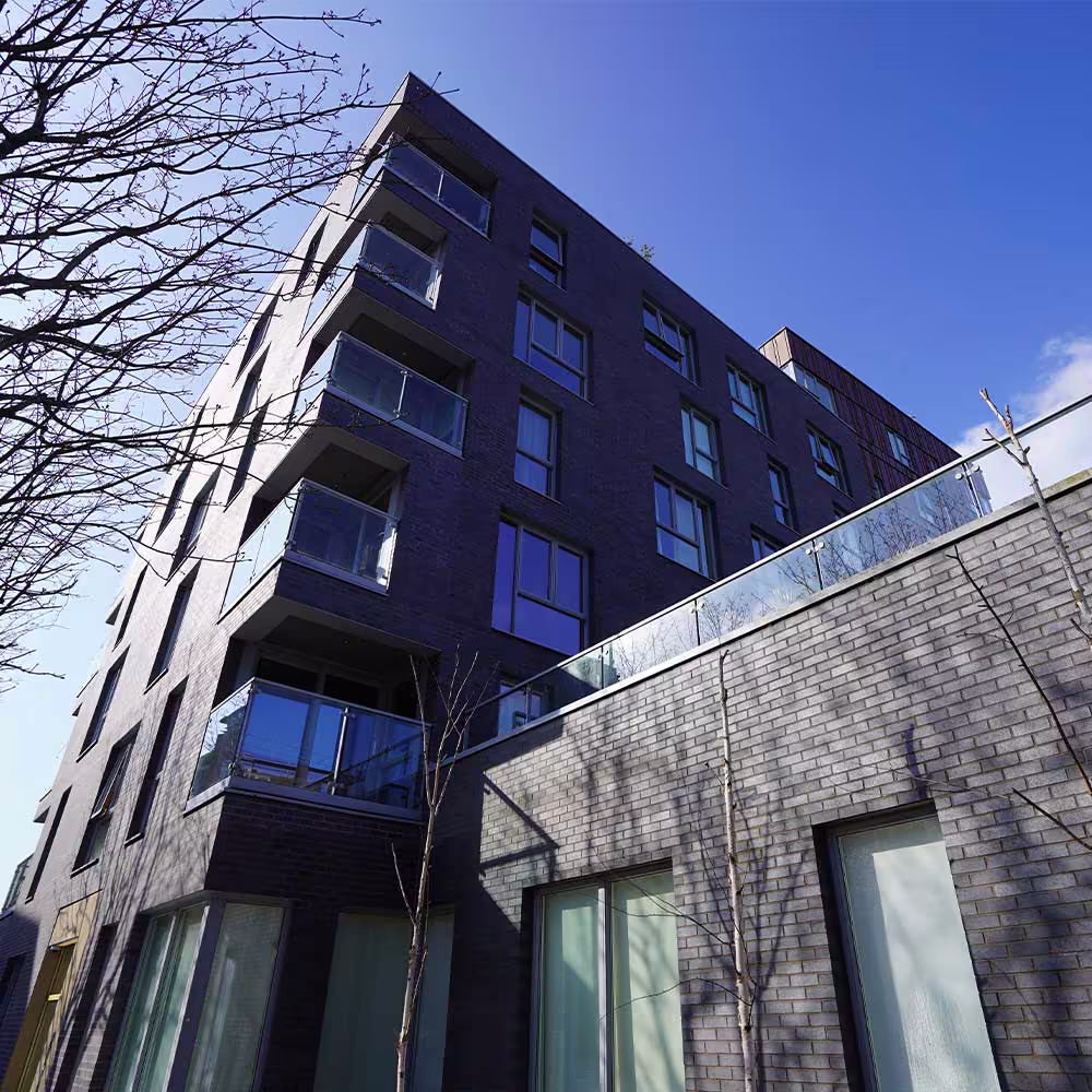 A low-angle perspective of the modern, dark grey brick apartment block, showing its clean lines and glass-fronted balconies against a clear blue sky.