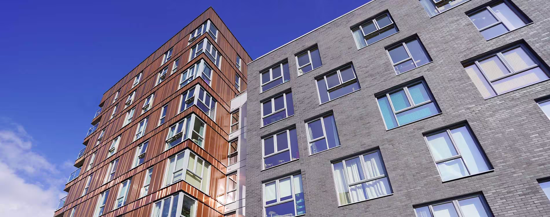 A low-angle view showing the contrast between two modern apartment blocks after a re-cladding project, one with striking copper-coloured metal cladding and the other with dark grey brick.