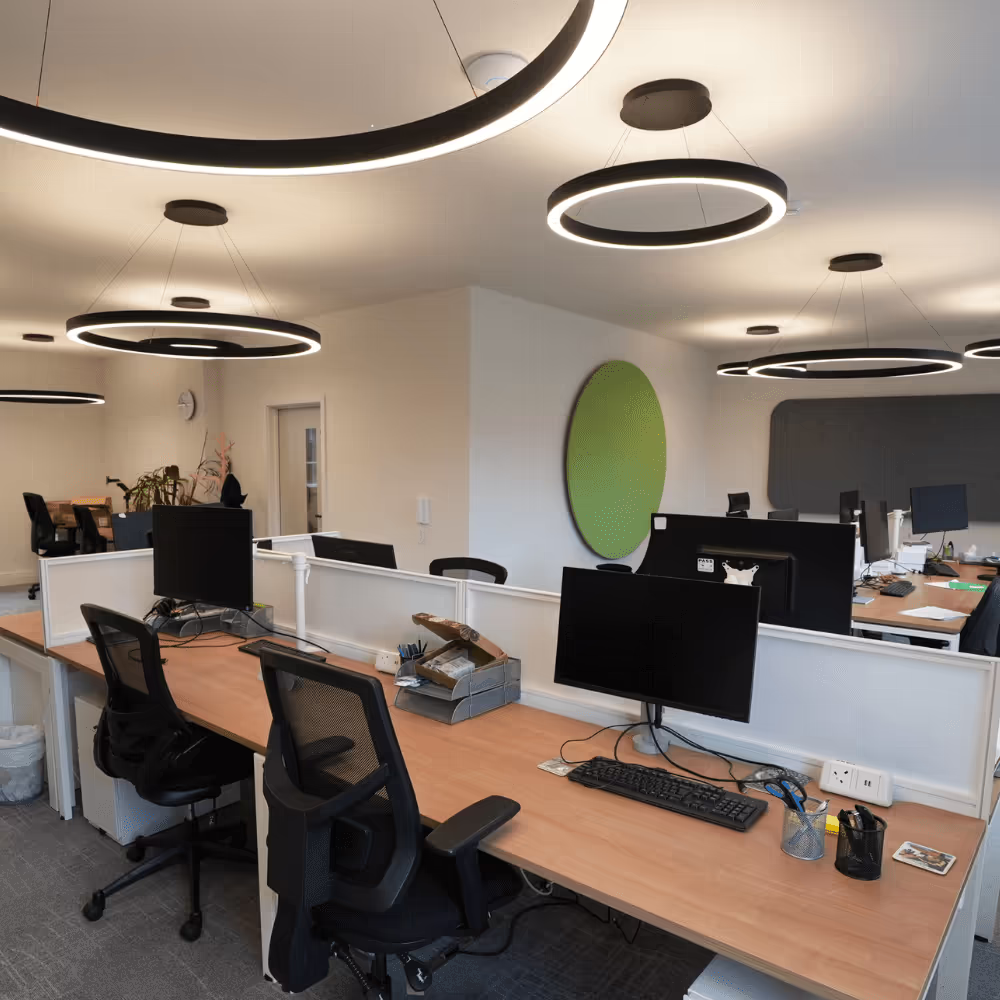 The remodelled open-plan office space, showing modern workstations with computers, ergonomic chairs, a green circular acoustic panel, and stylish circular LED lighting.