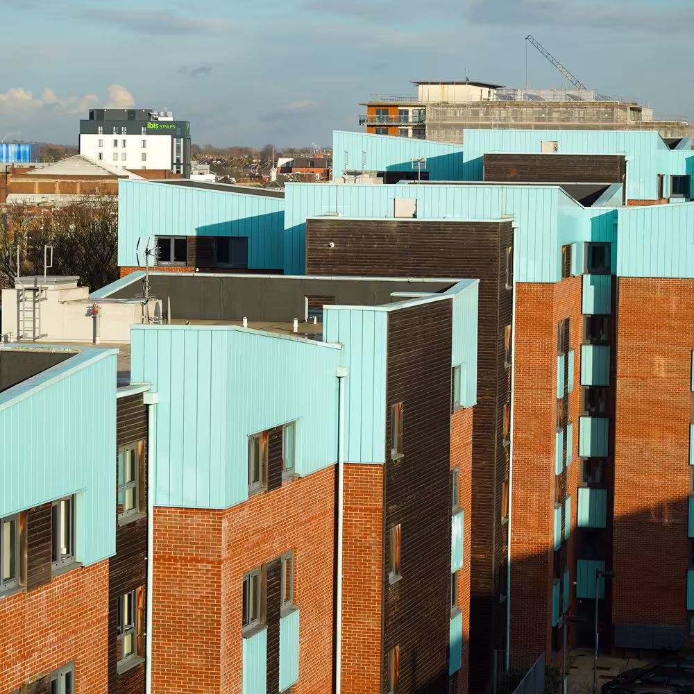 An aerial view over the rooftops of modern NHS keyworker accommodation, showing the unique architectural design of brick, wood, and turquoise metal cladding.