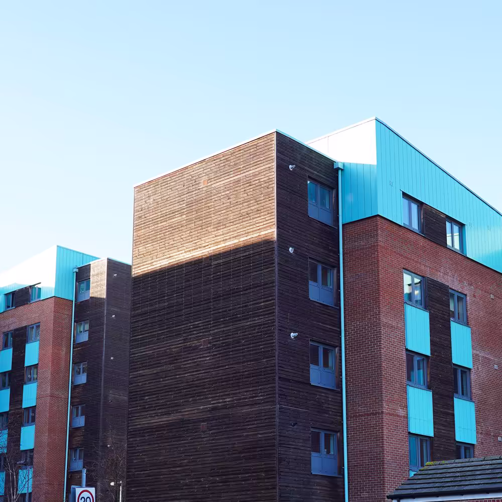 A low-angle architectural view of modern NHS keyworker accommodation buildings, highlighting the striking contrast between dark wood cladding and red brick with turquoise panels.