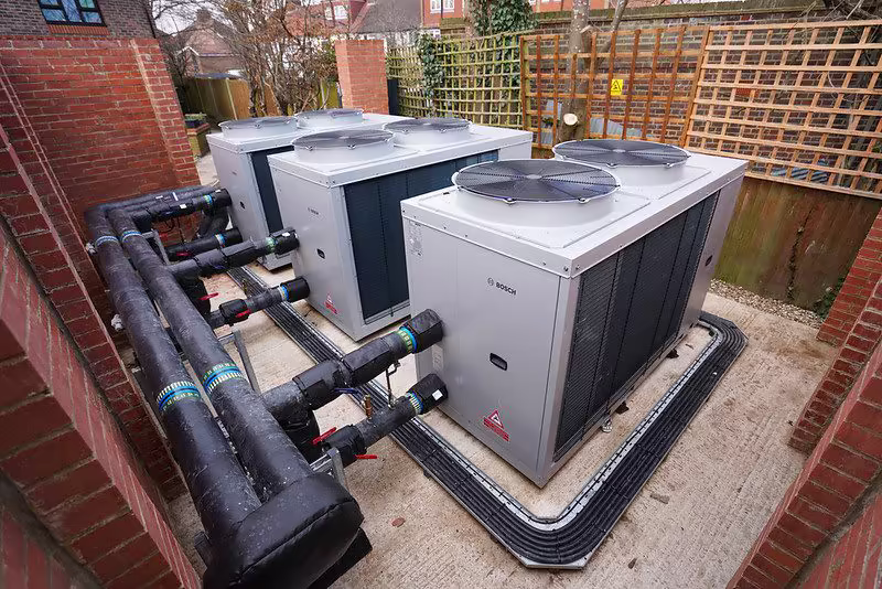 Four large Bosch commercial Air Source Heat Pump units installed outdoors, connected by thick, black insulated pipework to supply the new heating system.