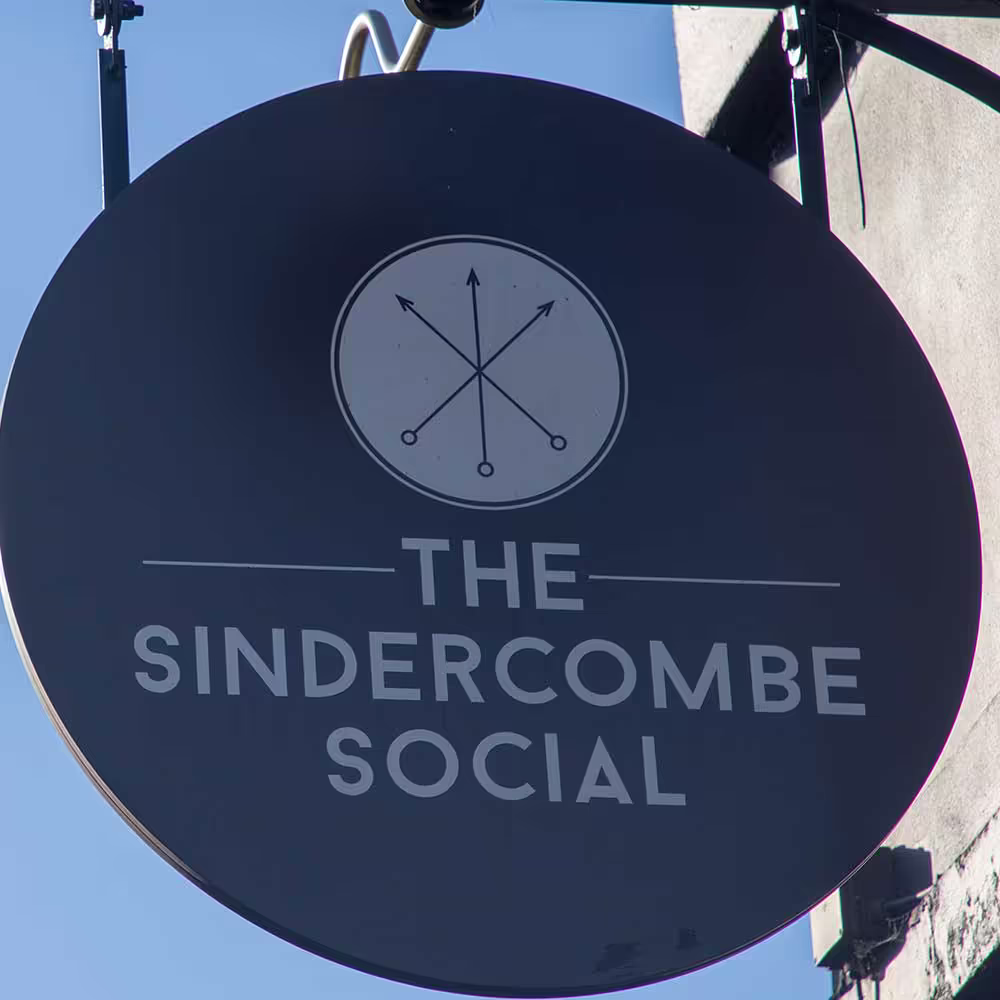 Close-up of the circular, dark navy exterior hanging sign for 'The Sindercombe Social', featuring the pub's name and compass-like logo.