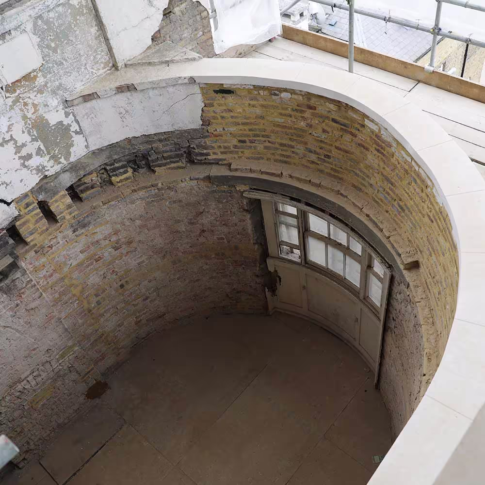 An overhead view showing a curved, cylindrical room space during building restoration. The walls are exposed, mixed-colour brick, and a curved, white stone balustrade or trim is being installed at the top.