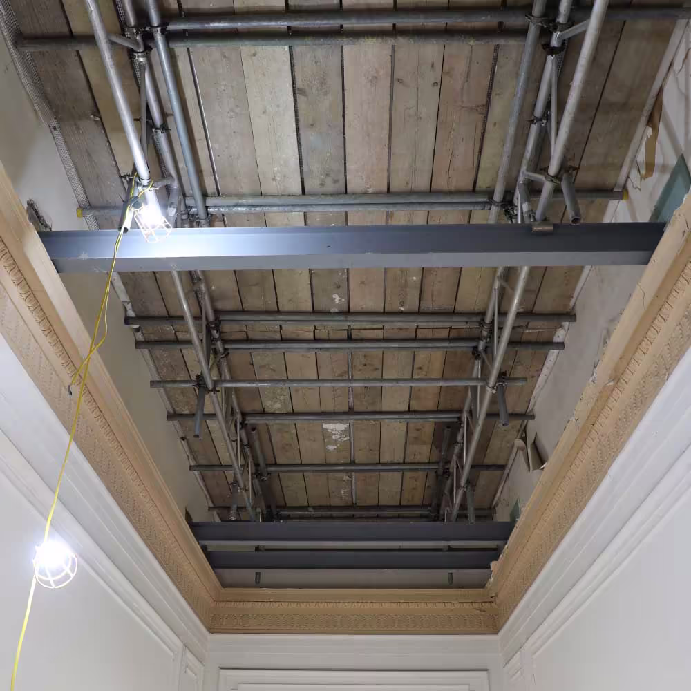 An upward-facing view of an ornate, white ceiling space with decorative cornice. Scaffolding is erected overhead, covering the ceiling, indicating restoration or repair work on the upper floor or roof structure.