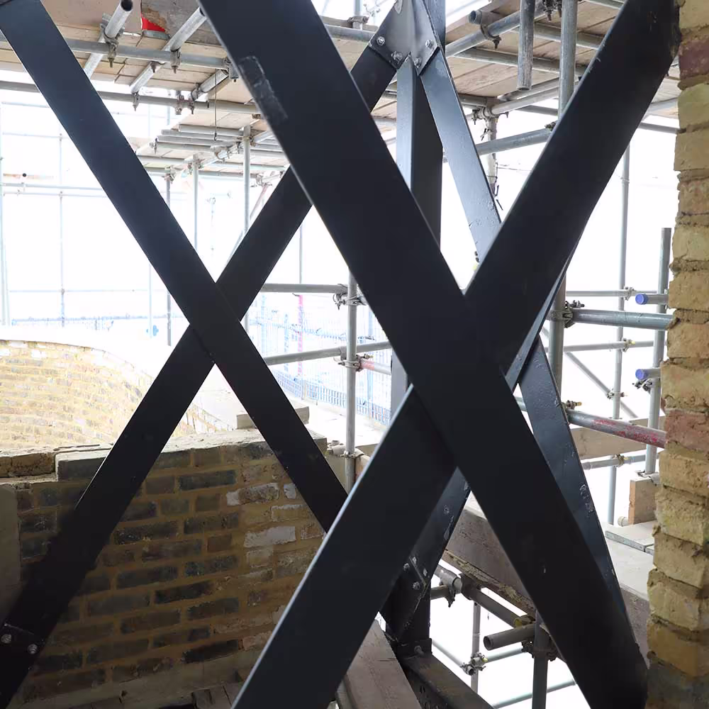 A structural detail shot showing black metal cross-bracing framing the view of extensive construction scaffolding in the background, set against exposed brickwork.