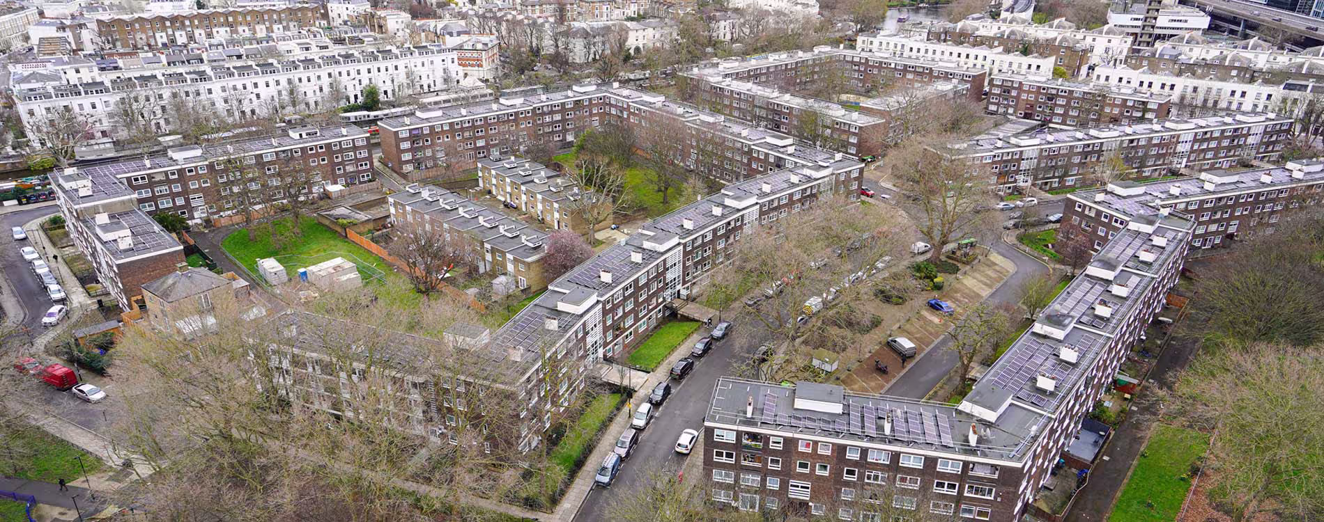 An aerial view of a large residential estate in a city, showing multiple low-rise apartment blocks arranged in a square and U-shape. Many of the building rooftops have visible arrays of new solar PV panels installed as part of energy retrofit works.