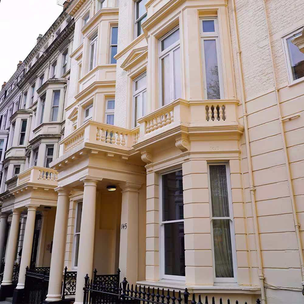 An angled view of the exterior of a refurbished pale yellow/cream townhouse, showing the entrance with columns, a bay window with a balustrade balcony, and the new, brightly framed windows from a roof and window repairs project.