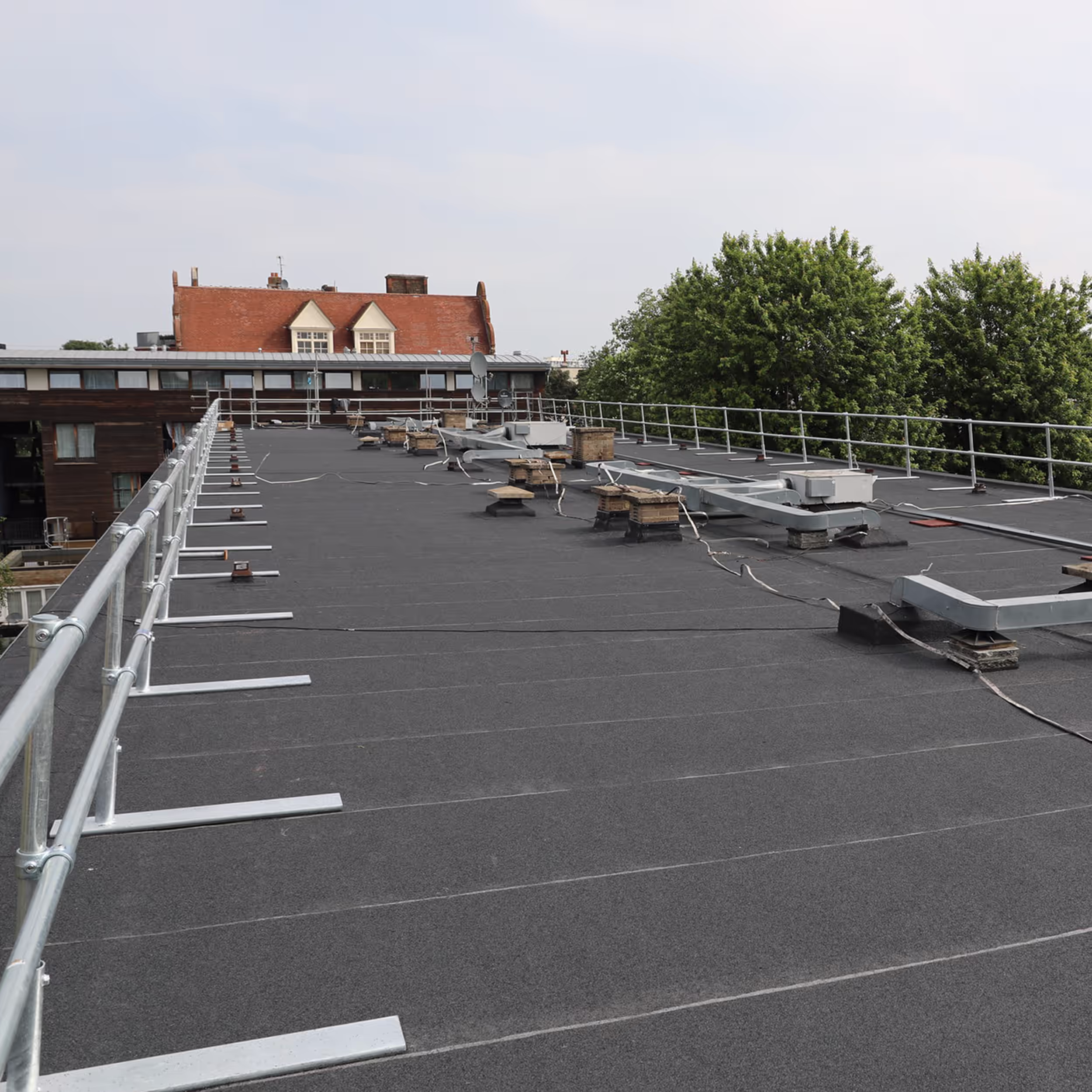 An angled view showcasing the full length of the newly installed non-penetrating metal safety railing (guardrail) along the perimeter of the renewed flat roof, with neighboring buildings in the distance.