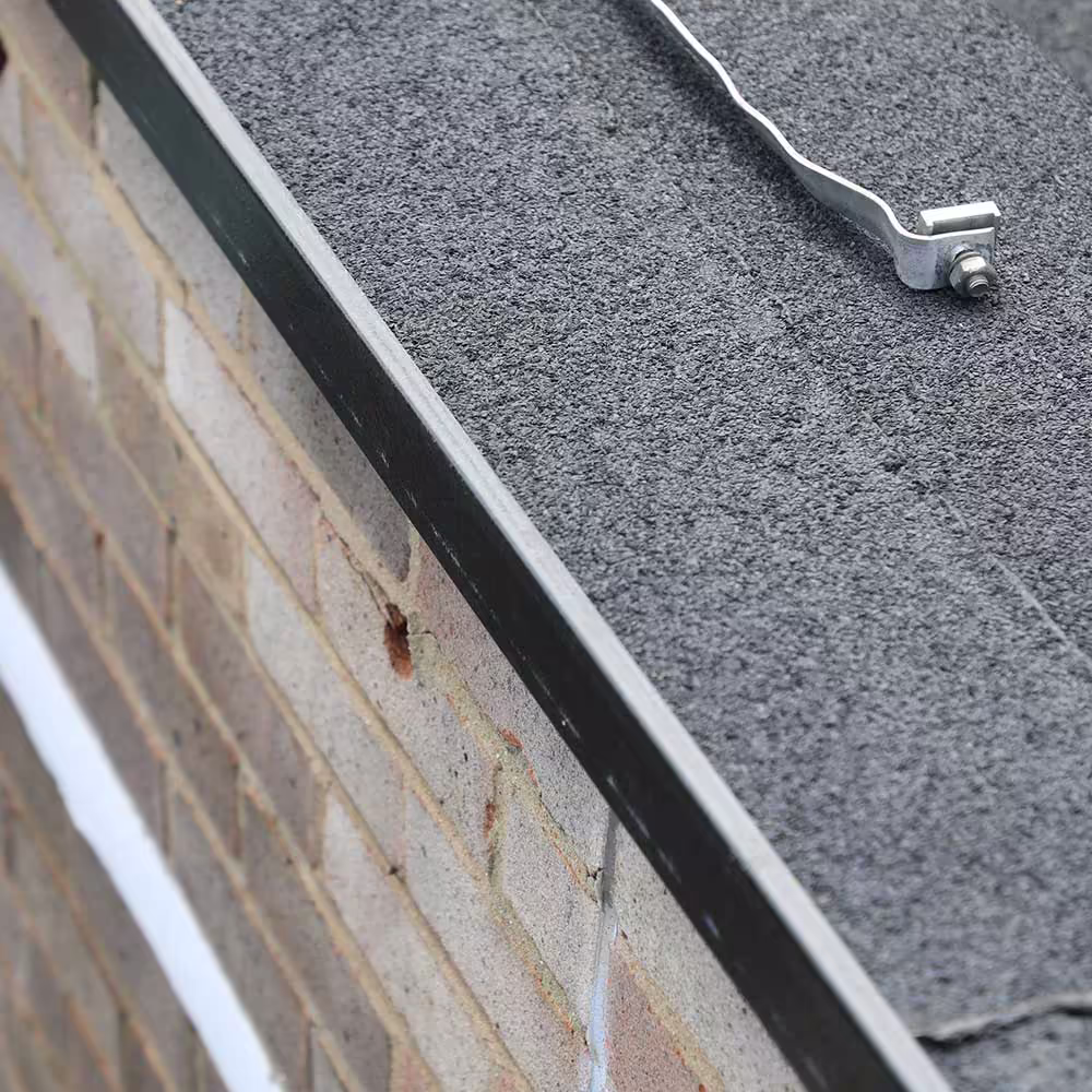 A close-up of the roof edge where the new flat roof membrane meets the brick parapet wall, showing the metal flashing and the granular surface of the roofing material, as well as a lightning protection conductor clip.