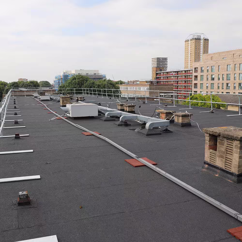 A view across the renewed flat roof, showing various ventilation ducts, chimney flues, and an array of non-penetrating guardrails installed at the roof edge for safety. The city environment is visible beyond the railing.
