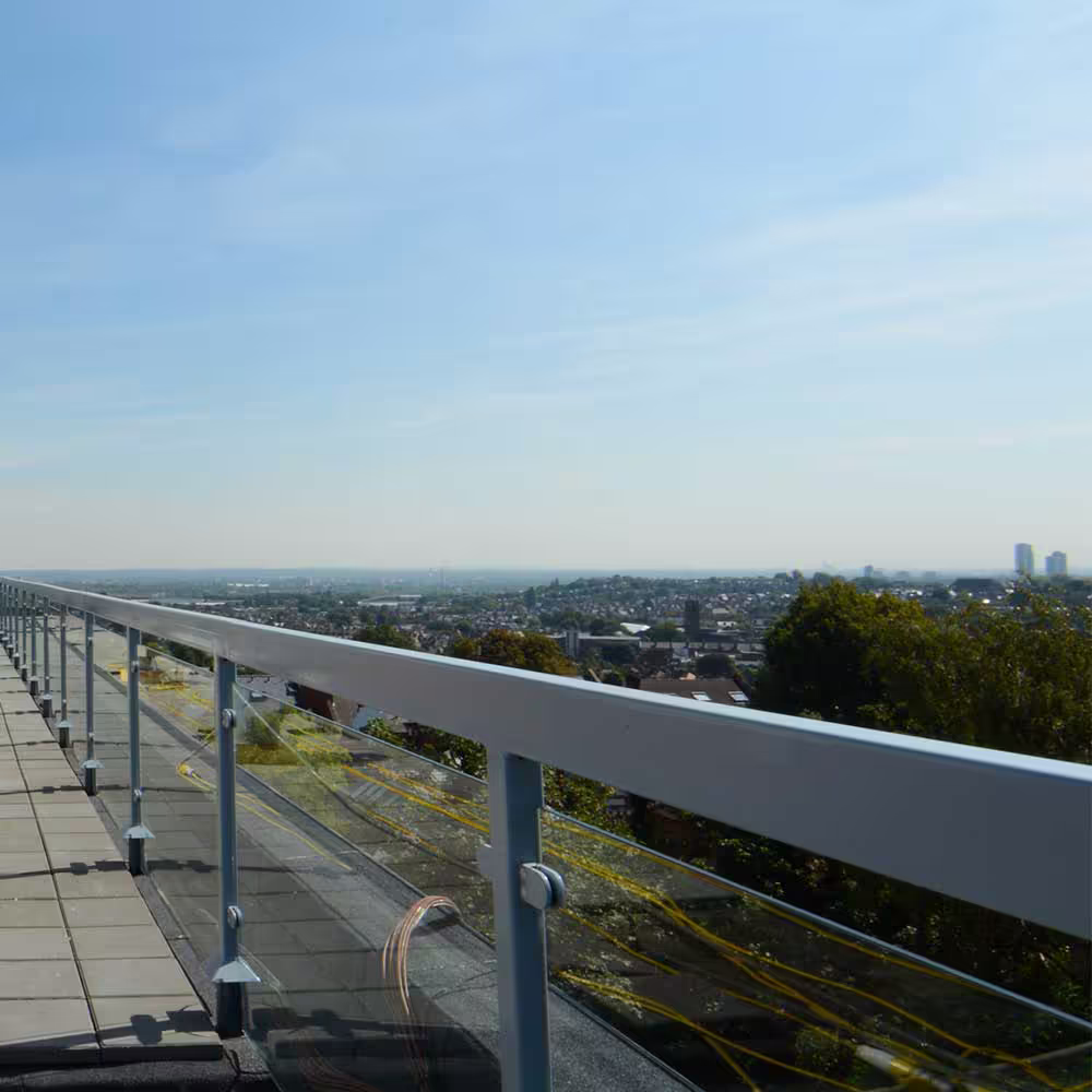A long-shot perspective along the newly installed safety railing of the rooftop walkway, showing the expansive view of the surrounding urban area under a bright blue sky.