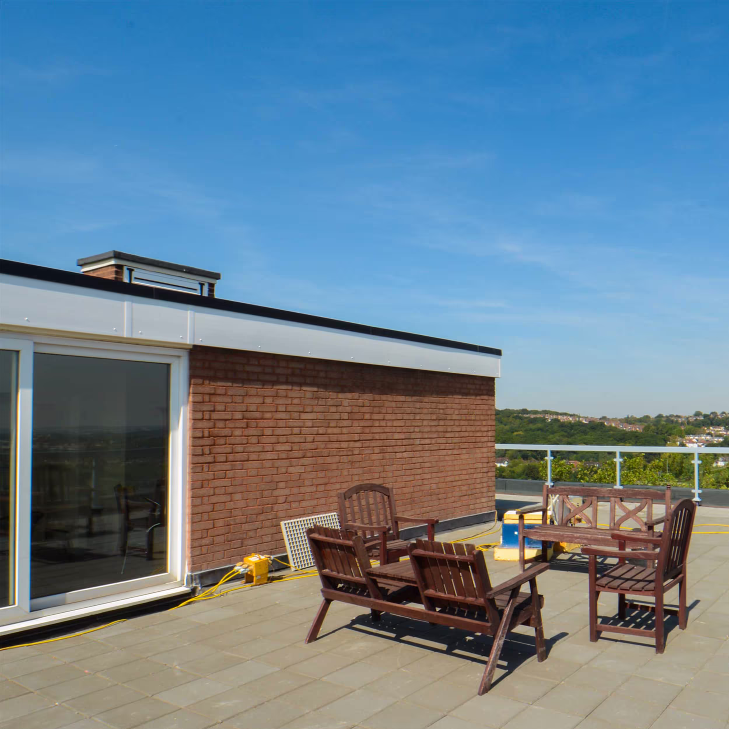 A section of the rooftop terrace with red brick walls, a large sliding glass door, and wooden patio furniture, indicating the completion of the rooftop repairs and conversion into a residential amenity.