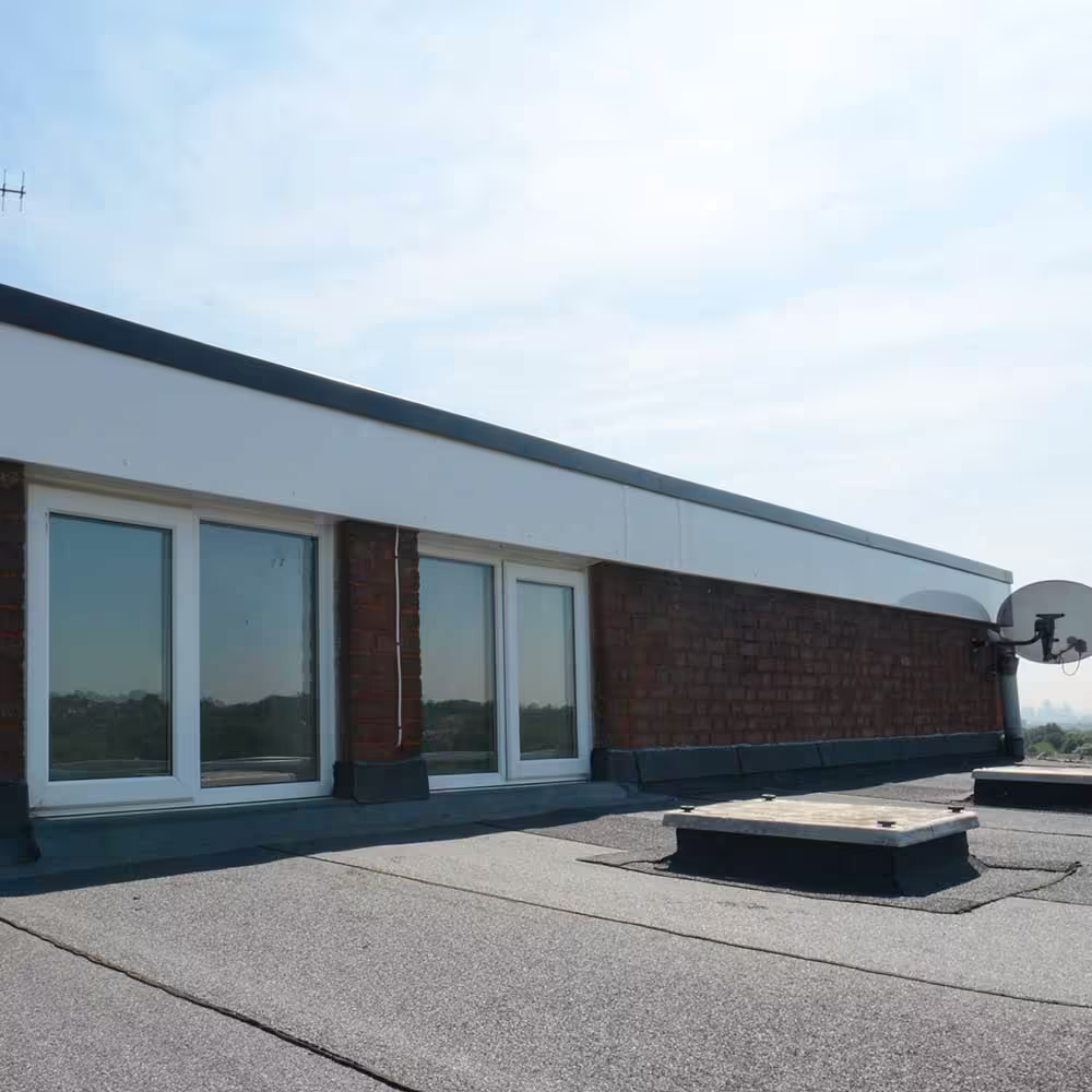 A close-up of a dark, granular flat roof surface with a section of red brick parapet wall, new windows, and installed skylight hatches, showcasing the quality of the rooftop repairs and maintenance.