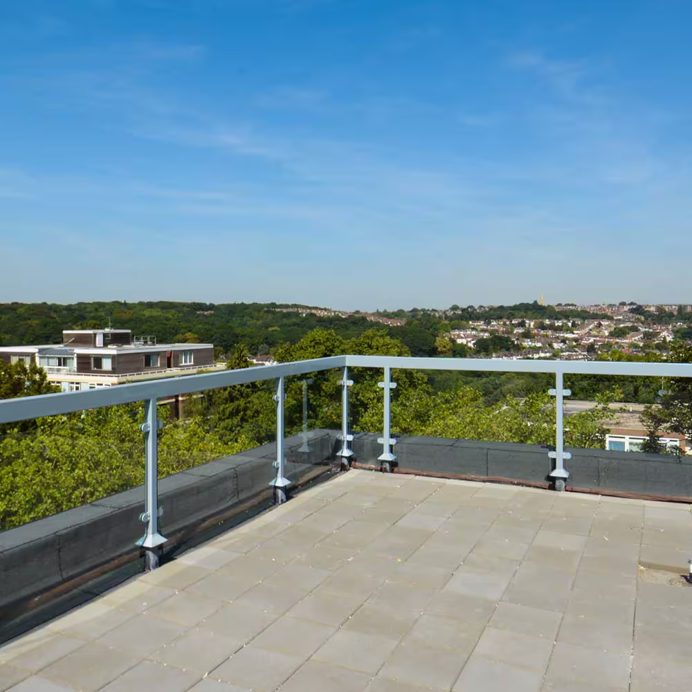 A corner view of the newly paved rooftop terrace, enclosed by a clean, modern glass and metal railing, overlooking a lush, green urban landscape and distant housing, demonstrating the finished rooftop maintenance.