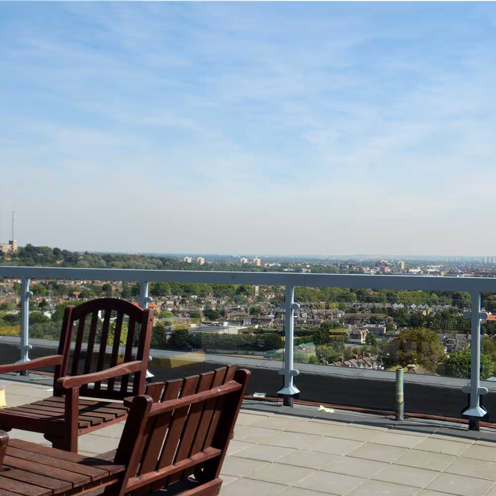 Wooden garden chairs are set up on the rooftop terrace, offering a scenic view over a green residential area and distant city skyline, highlighting the transformation into a usable rooftop amenity space.