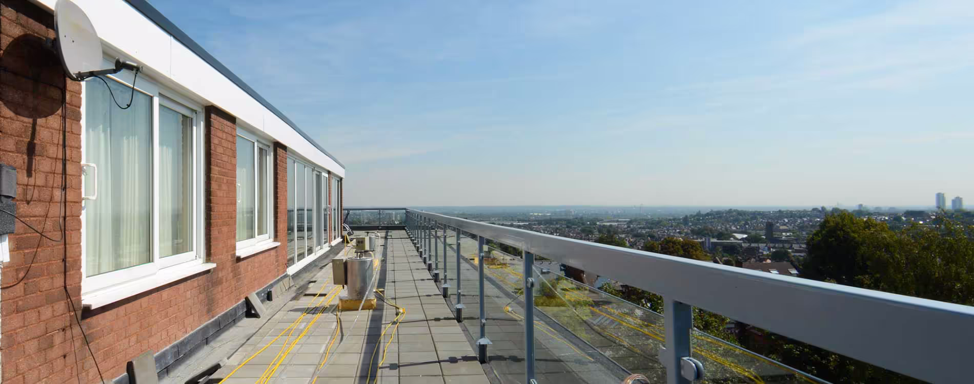A wide, elevated view of a refurbished rooftop terrace featuring a paved surface, modern glass and metal guardrails, and a panoramic city skyline in the distance. The long row of upper-floor windows and red brick wall are visible on the left.