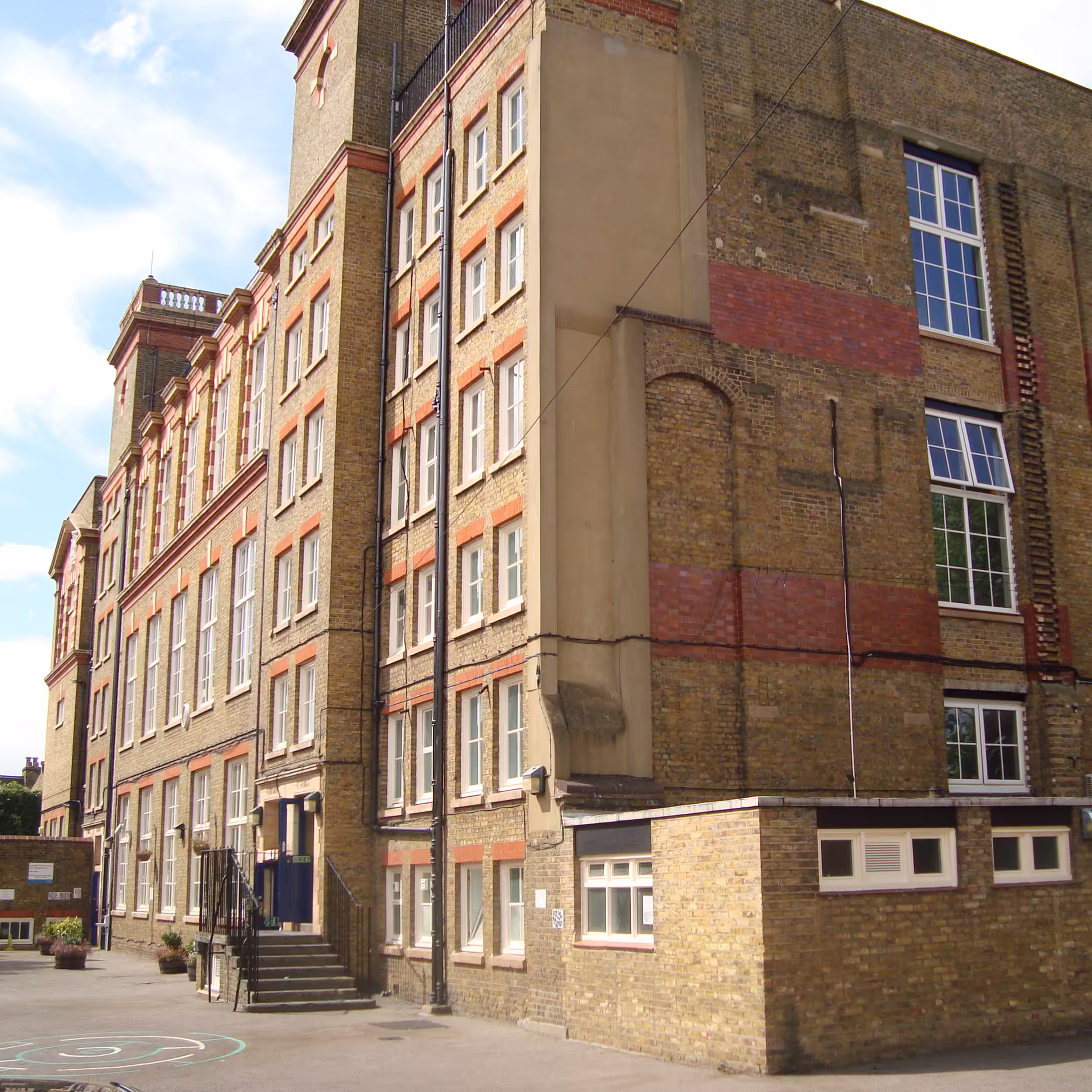 A full side view of the large, multi-story Victorian-era school building, featuring long, vertical columns of white-framed windows and a small staircase entrance to the playground.