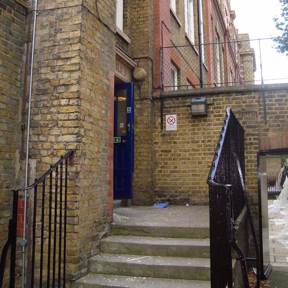 A close-up of a restored brick entrance corner of the school building, showing concrete steps with a black metal handrail leading up to a blue door. The distressed brickwork suggests historical building renovation.