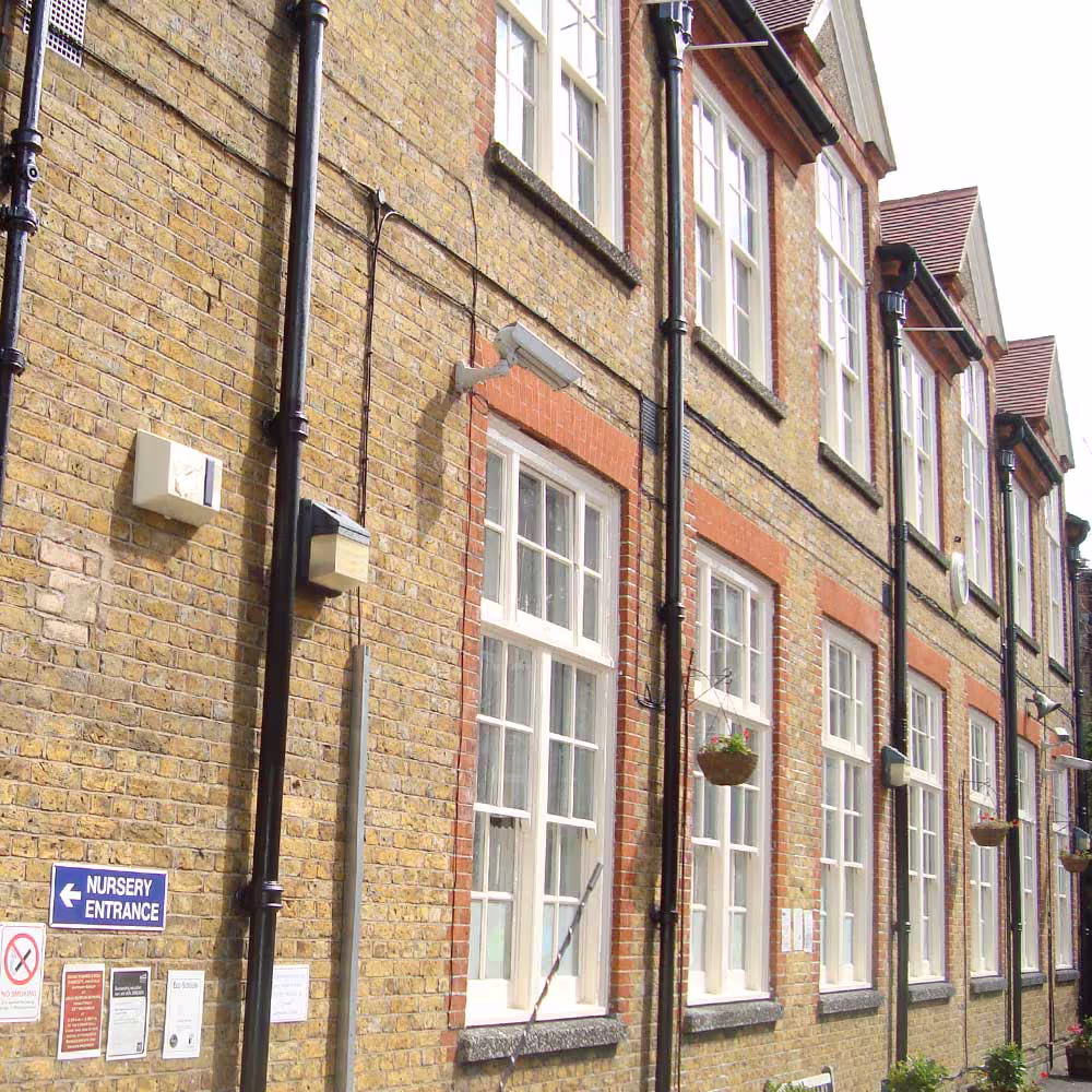 A close-up of a section of the renovated school's yellow brick wall, showing black external piping, utility boxes, a 'Nursery Entrance' sign, and a row of large, white-framed windows with red brick sills.