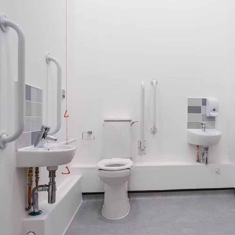 A clean, newly renovated accessible washroom featuring a toilet, low sink, and prominently installed white grab bars for support, with a gray tiled wall and safety cord, demonstrating ADA-compliant renovations.