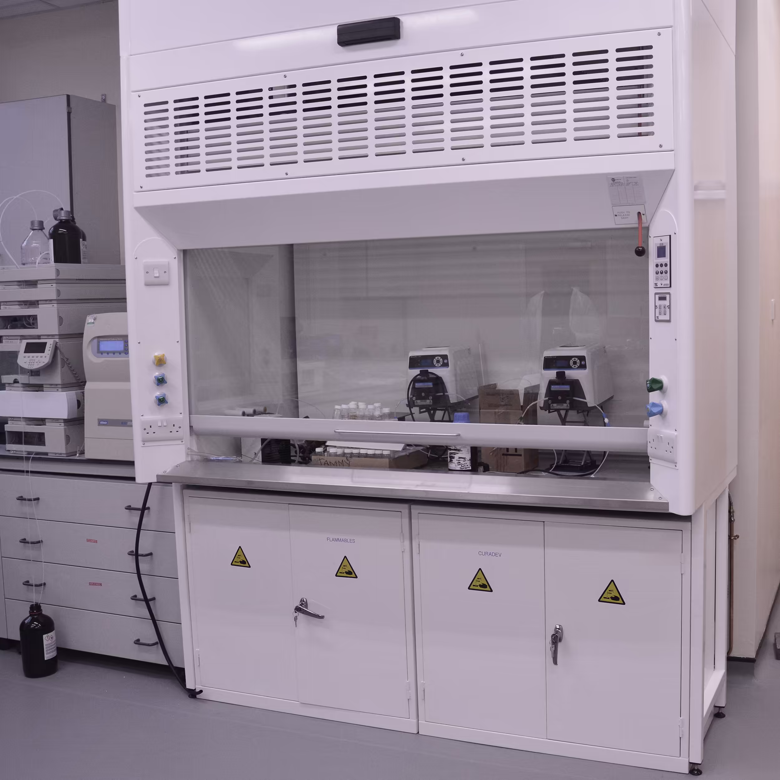 A detailed shot of a new white fume cupboard in a refurbished science lab. The unit includes locked cabinets labeled 'FLAMMABLES' and 'CUPBOARD', a glass sash, and control panels for ventilation, showing professional laboratory installation.