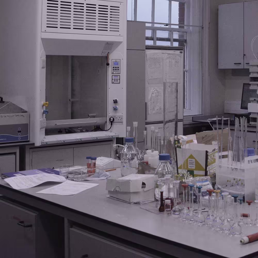 A close-up of a workbench in the science lab with various chemistry glassware, including test tubes, flasks, and beakers, next to a fume cupboard, illustrating the functional lab environment.