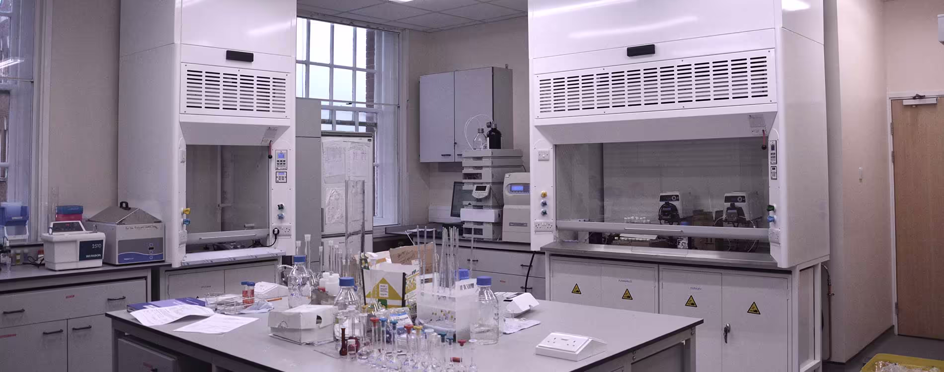 A wide view of a modern, refurbished science laboratory, featuring two large white fume cupboards, grey workbenches cluttered with glassware and beakers, and analytical equipment near a window, showcasing the completed lab fit-out.