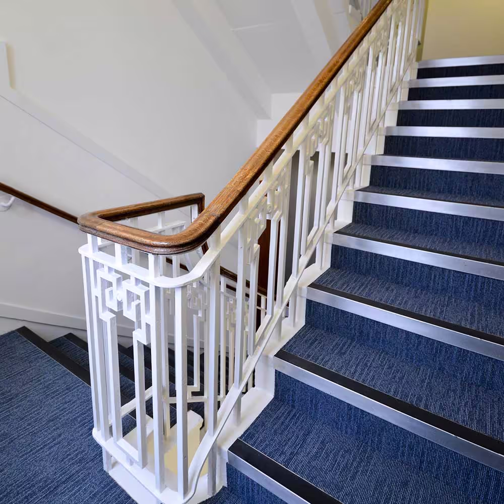 A close-up of a modernized office staircase, featuring white decorative metal balusters with a polished wooden handrail and steps covered in dark blue patterned carpet, indicating a stairwell refurbishment.