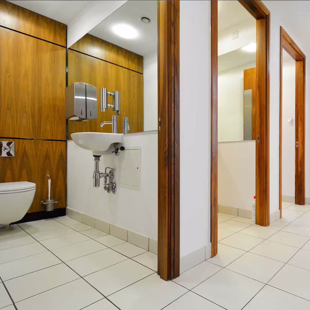 A renovated modern washroom interior, featuring white tiled floors, a wall-mounted sink and toilet, and warm, horizontally paneled wooden walls around a mirror, with light wood door frames leading to adjacent rooms.