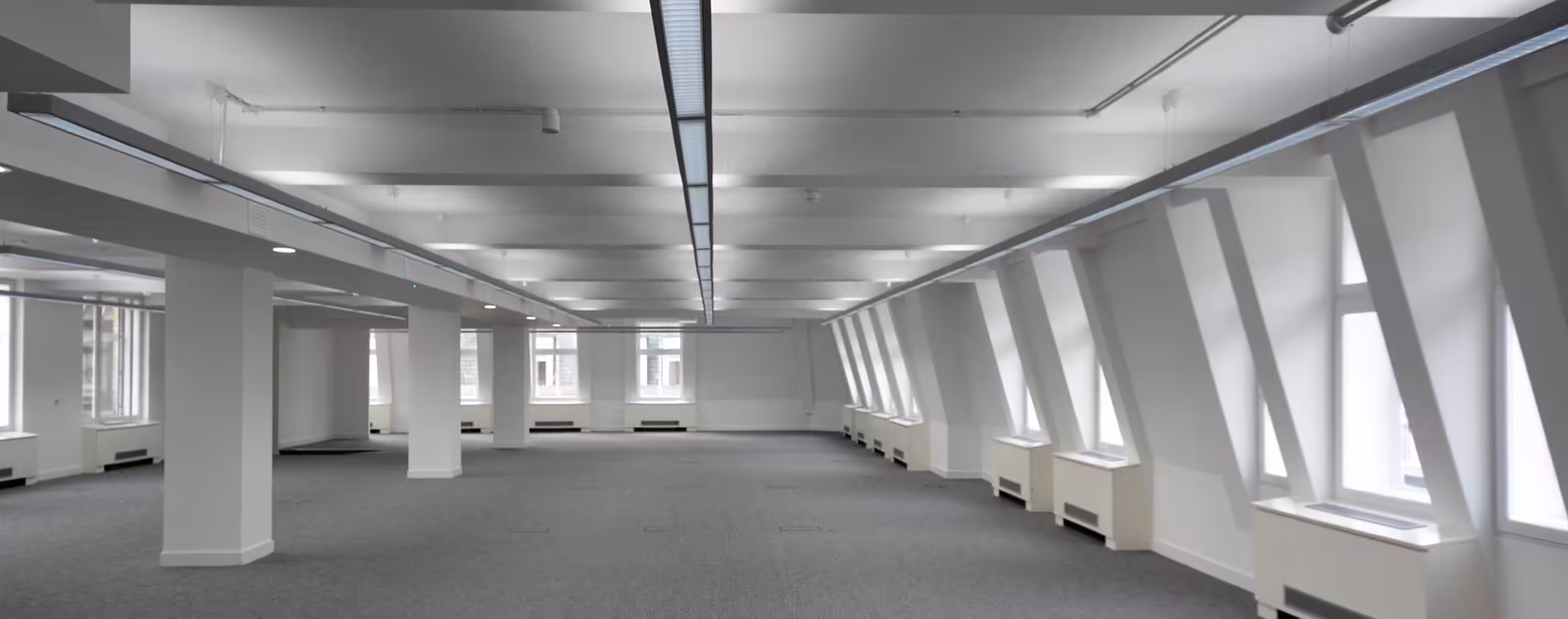 A wide, interior view of a newly stripped-out commercial office space. The room features a long central light fixture, white structural columns, and distinctive angled window bays, showcasing the empty canvas ready for fit-out works.