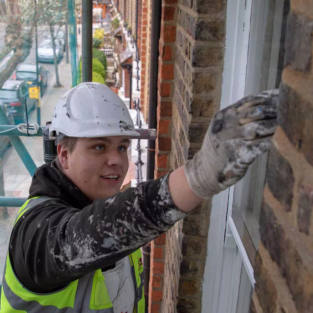A young male Axis tradesperson on scaffolding, wearing a hard hat and high-visibility vest, paints a white window frame next to a brown brick wall on a busy street, highlighting external property maintenance.