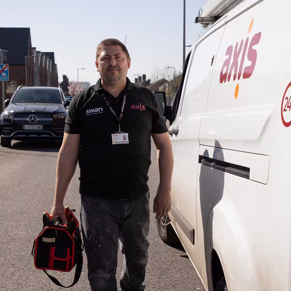 A male Axis technician in a black uniform and jeans walks away from a branded white van, carrying a red and black tool bag, ready to begin work on a sunny residential street.