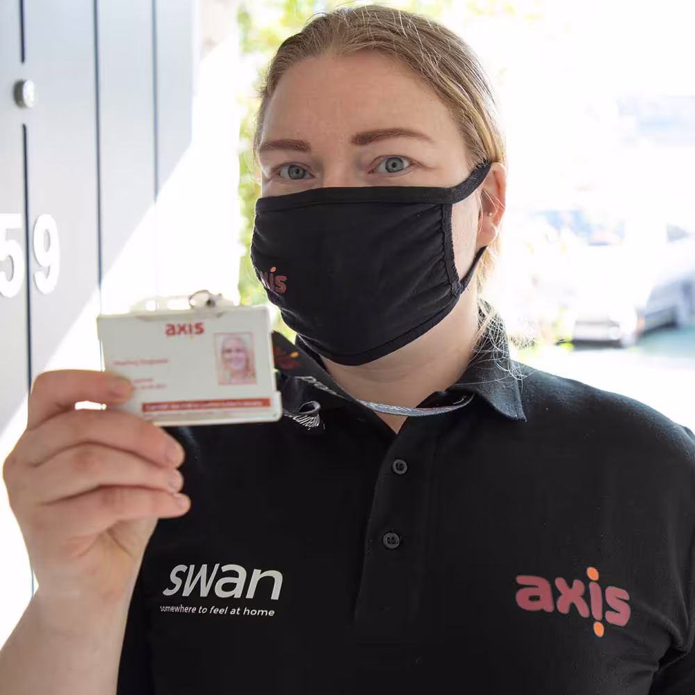 A female Axis employee in a black uniform and face mask holds up her ID badge with the 'Axis' logo, assuring safety and professionalism for a property management service.
