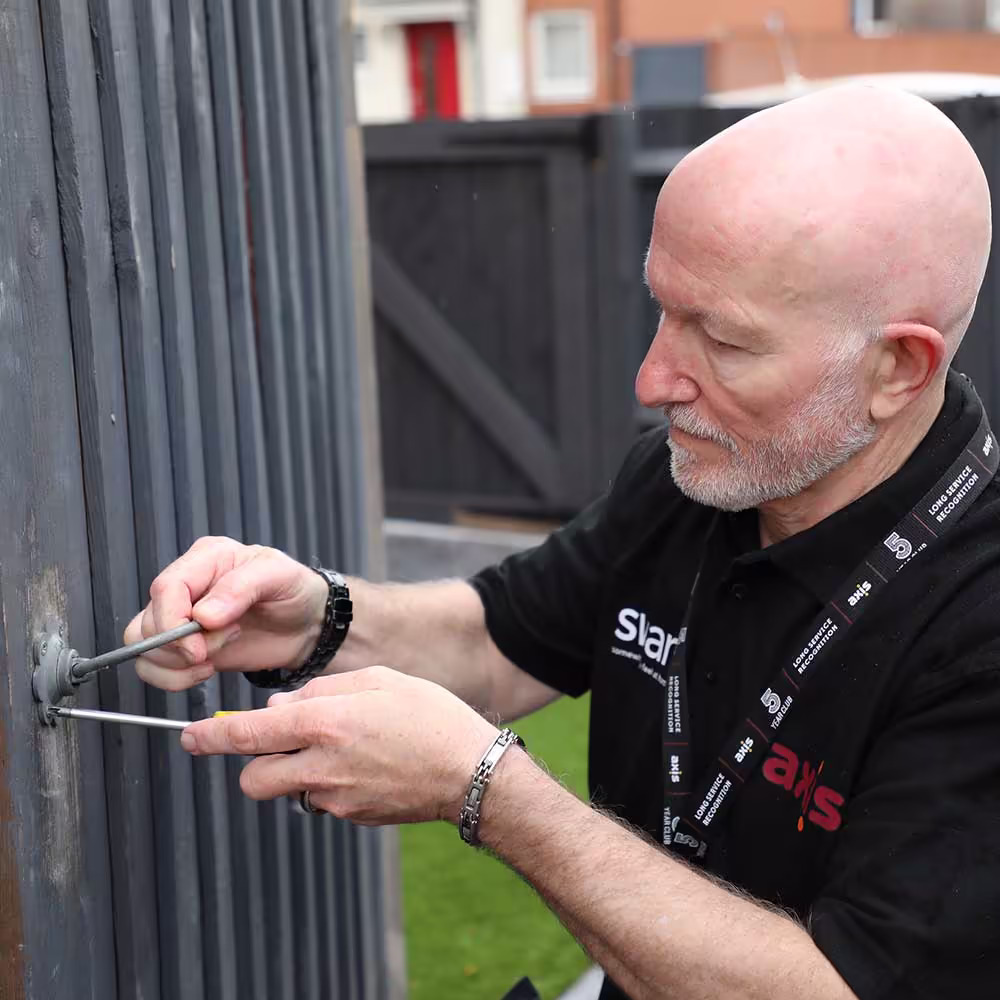 A close-up of a bald male technician in a black Axis uniform and lanyard, with a wristwatch, using screwdrivers to work on the latch or lock of a dark grey wooden fence panel, performing fencing or gate repair and maintenance.