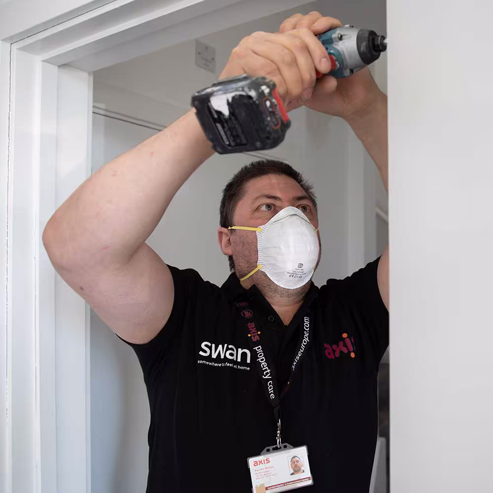 A male Axis technician in a black uniform and respirator mask uses a cordless drill to install or repair a door frame or ceiling inside a property, emphasizing safety during internal property repair.