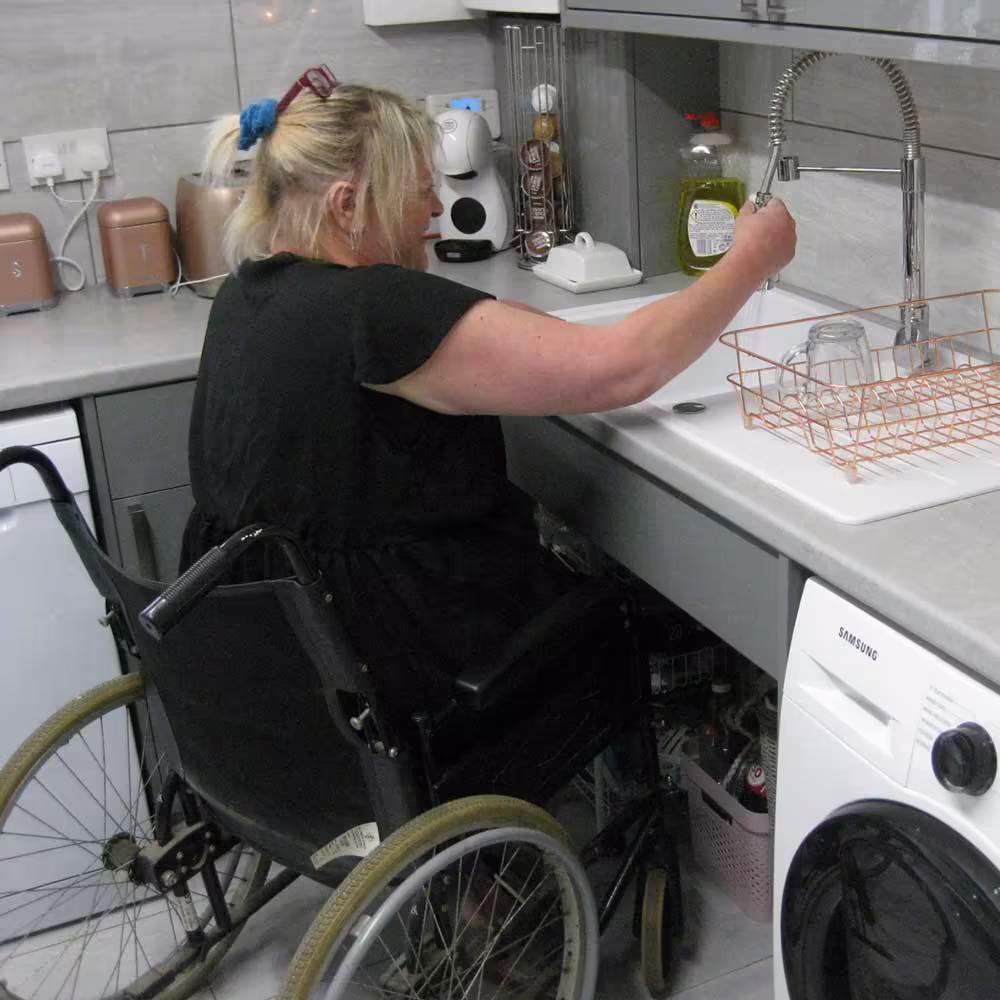 A woman in a wheelchair uses a height-adjustable sink or counter in a modern kitchen, demonstrating the successful installation of an adapted or accessible kitchen as part of a total asset management programme.