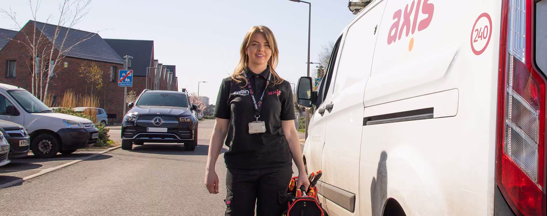 A smiling female Axis technician in a black uniform and lanyard walks away from a white branded service van, holding a red tool bag, on a street lined with modern houses, representing property asset management service.