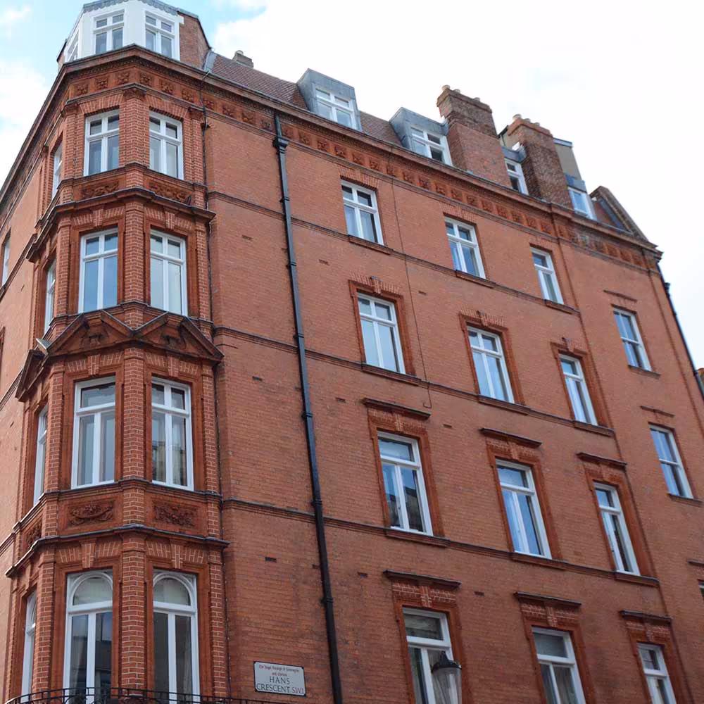 An upward-angled view of the corner of the renovated red brick Victorian apartment building, highlighting the tiered bay windows, decorative brickwork, and new white window frames below the dormer roofline.