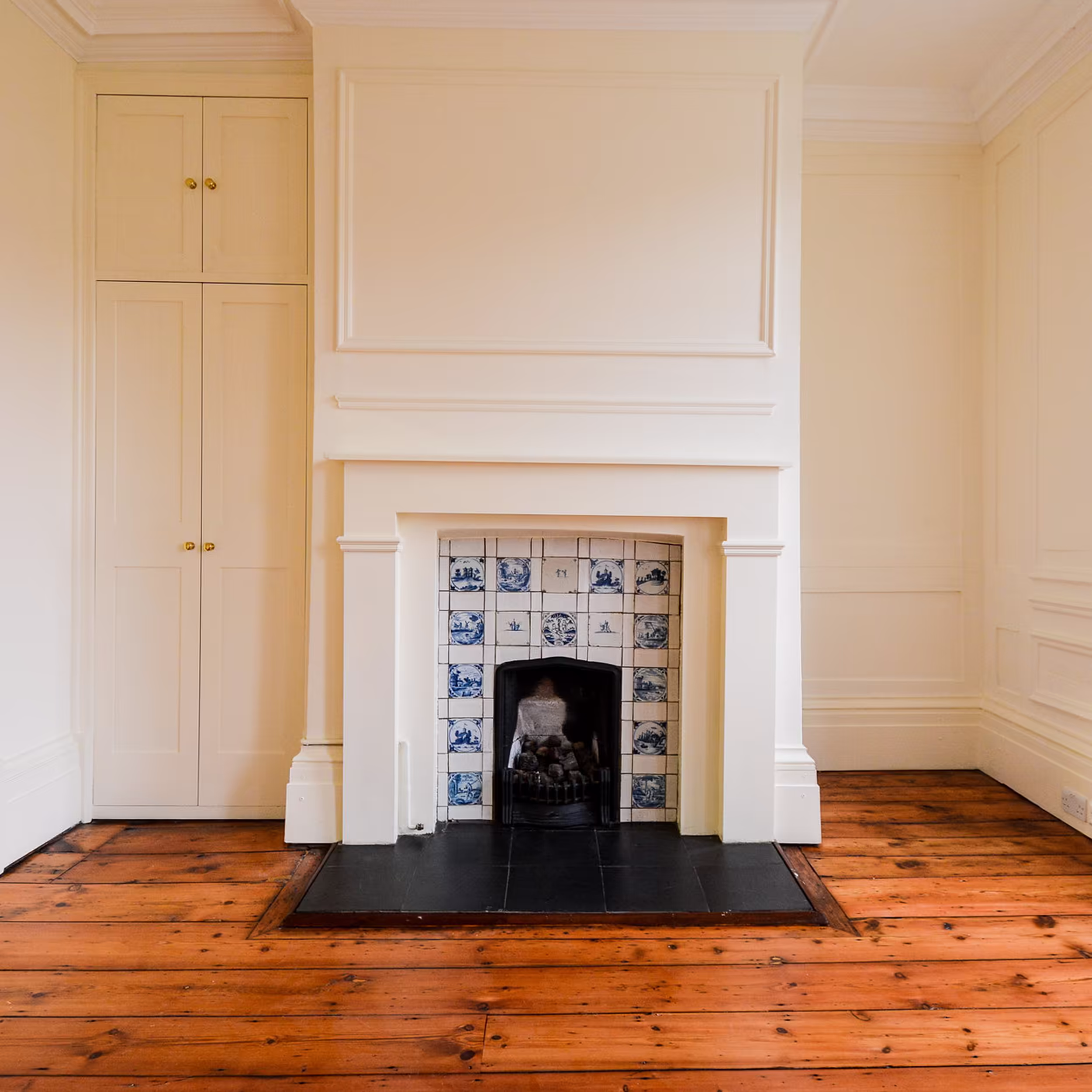 The interior of a refurbished room showing original architectural details, including a white painted fireplace mantel with blue and white Delft-style tiles, built-in cupboards, and polished wooden floorboards, ready after a void property refurbishment.