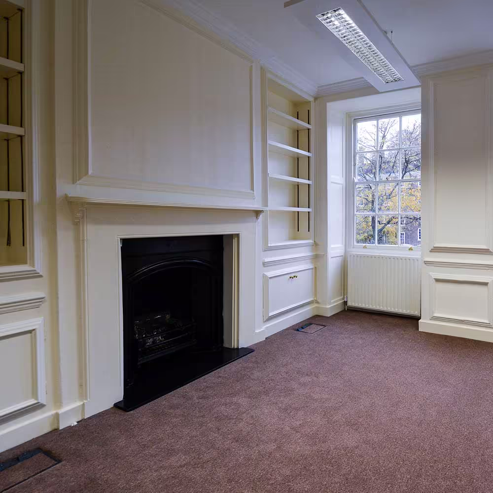 The interior of a renovated room with tan carpet, built-in shelving, an elegant white fireplace surround with a black iron grate, and a large white sash window that provides natural light.