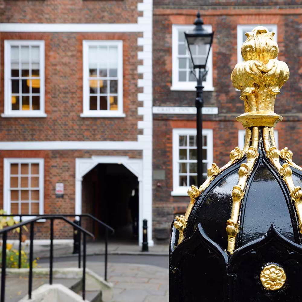 A close-up of an ornate black and gold metal post finial and a street lamp, set against the backdrop of the red brick building's entrance archway, showing the detailed exterior metalwork restoration.