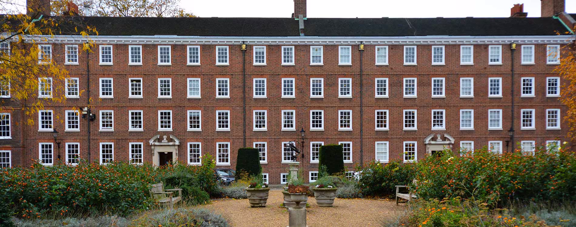 A wide, front-on view of a large, traditional red brick building with many uniform white sash windows and a dark roof, seen from across a manicured communal garden, showcasing the exterior of the refurbished property.