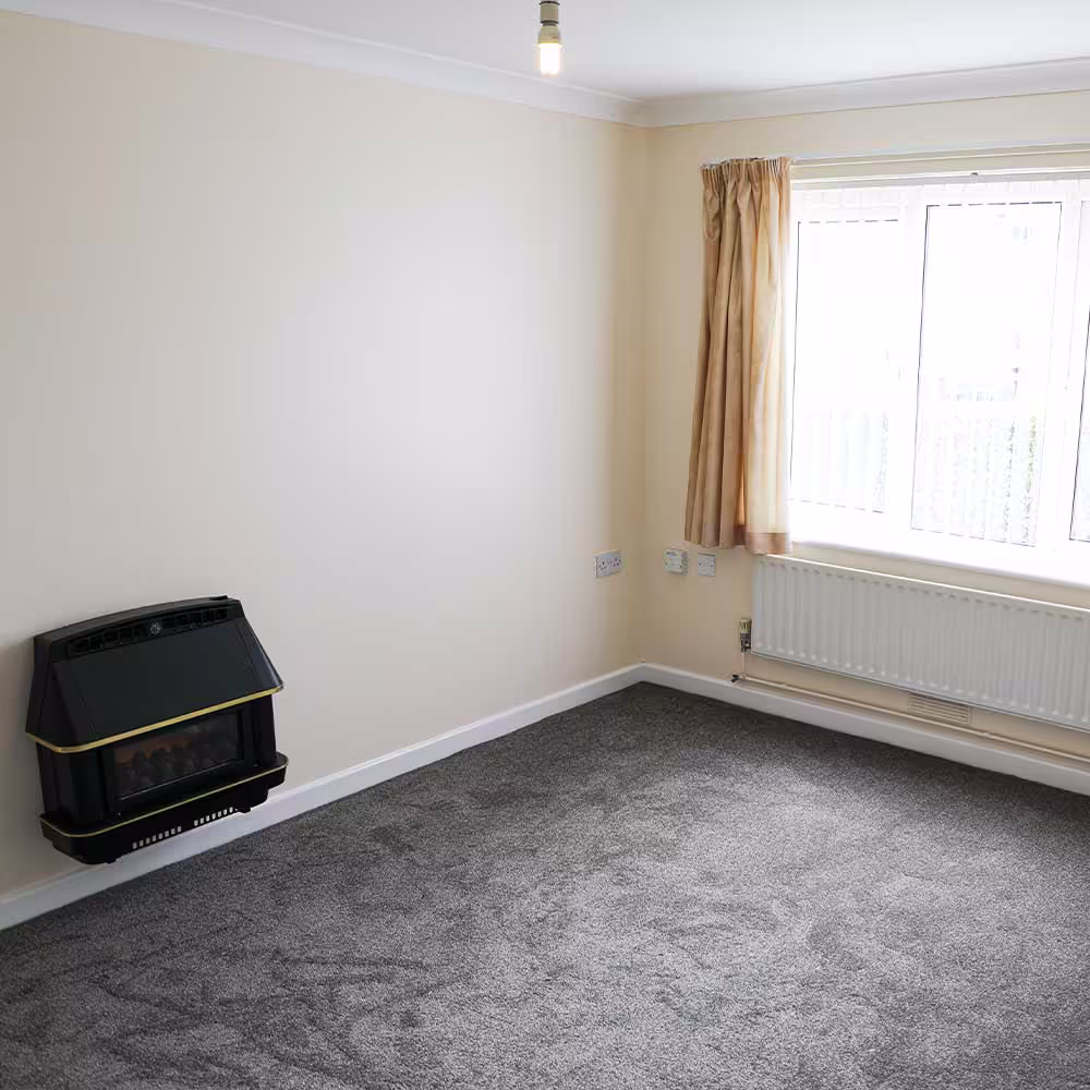 A clean, empty living room in a refurbished void property, featuring new grey carpet, cream-colored walls, a gas wall heater, a white radiator, and a window with beige curtains, illustrating a completed internal renovation.