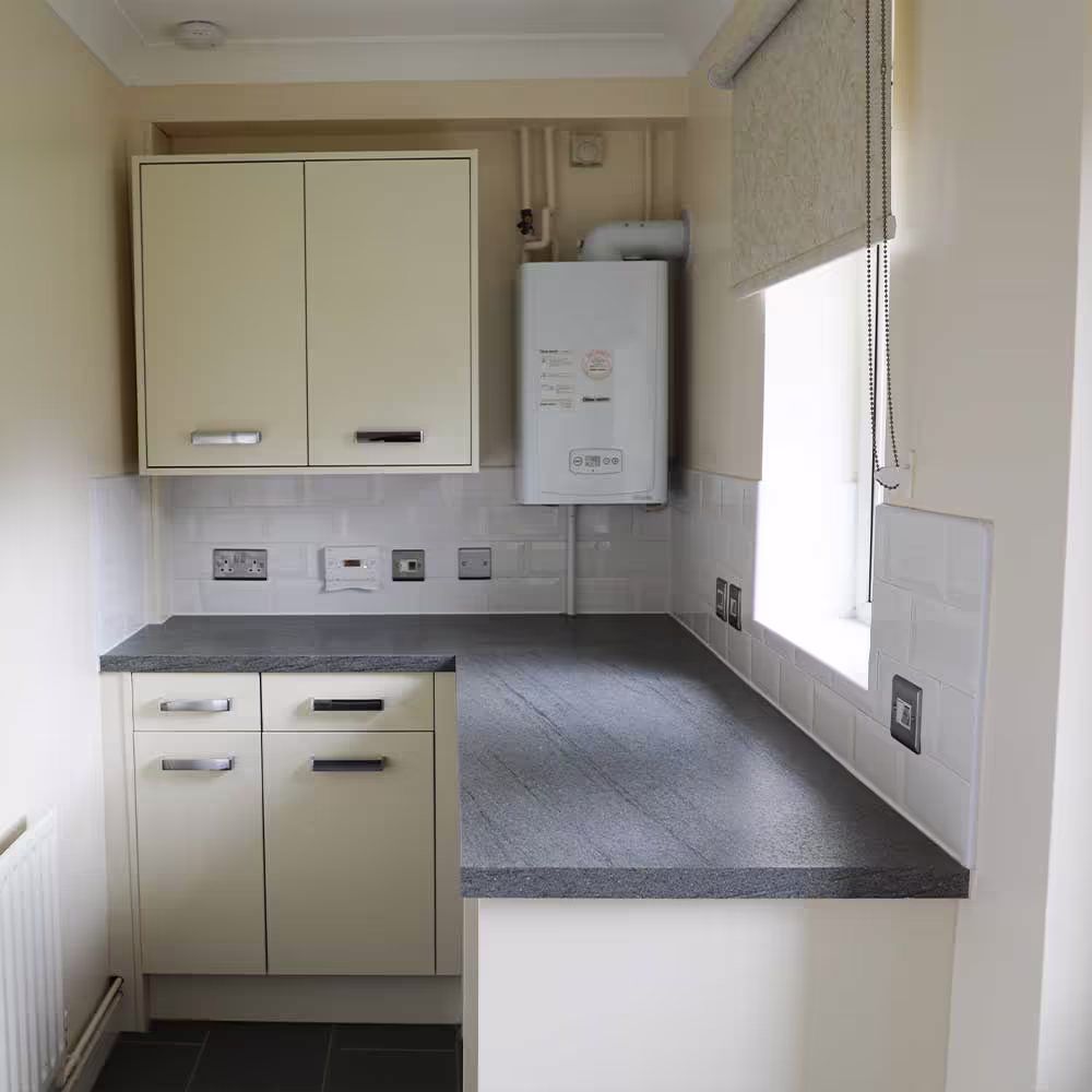 A newly installed utility area in a kitchen, featuring light cream cabinets, a dark grey countertop, white subway tile splashback, and a new wall-mounted gas boiler, showcasing a void property refurbishment.