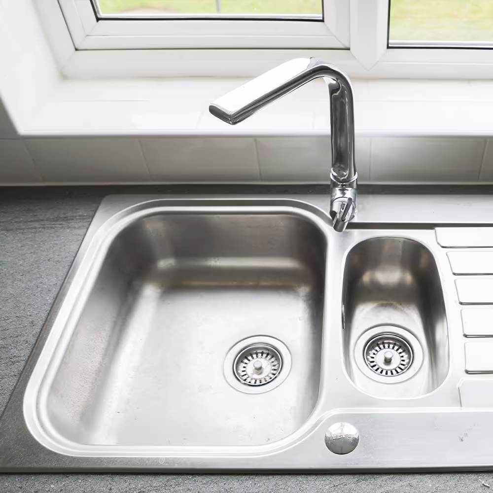 A close-up of a new stainless steel kitchen sink with a modern chrome swan-neck tap, set into a dark grey countertop by a window, highlighting the quality of the new kitchen fittings.